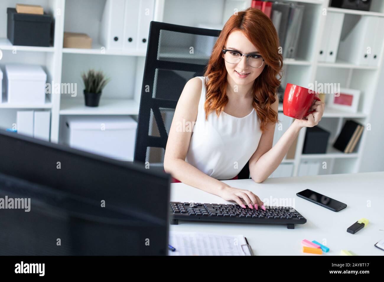Belle jeune fille est assise au bureau dans le bureau, tient une tasse dans une main et travaille à l'ordinateur. Banque D'Images