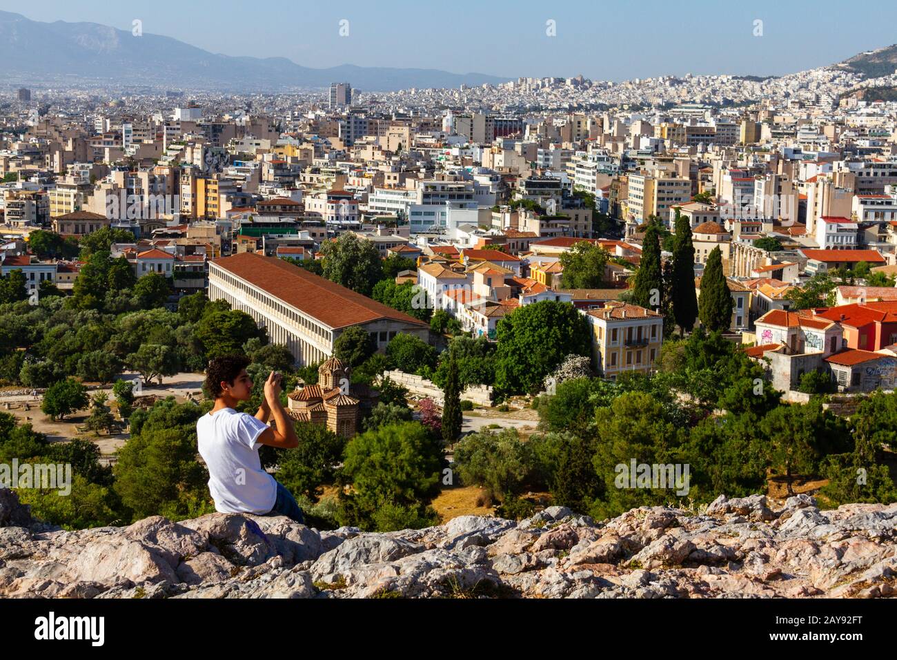 Toutiste assis sur un rocher à travers l'Acropole en prenant une photo du paysage urbain d'Athènes un après-midi d'été Banque D'Images