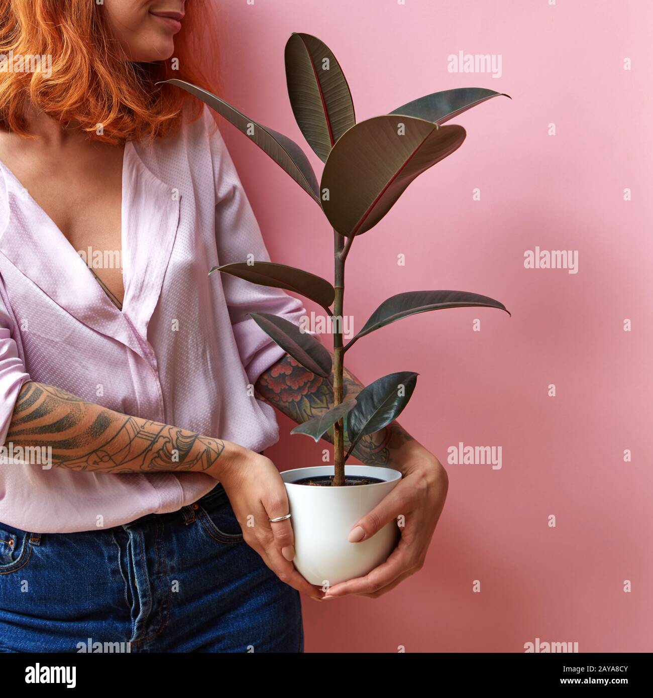 Sourire heureuse fille avec des cheveux de gingembre tient flowerpot avec ficus de plante verte, mains de tatouage sur un fond rose, espace de copie. Ca Banque D'Images