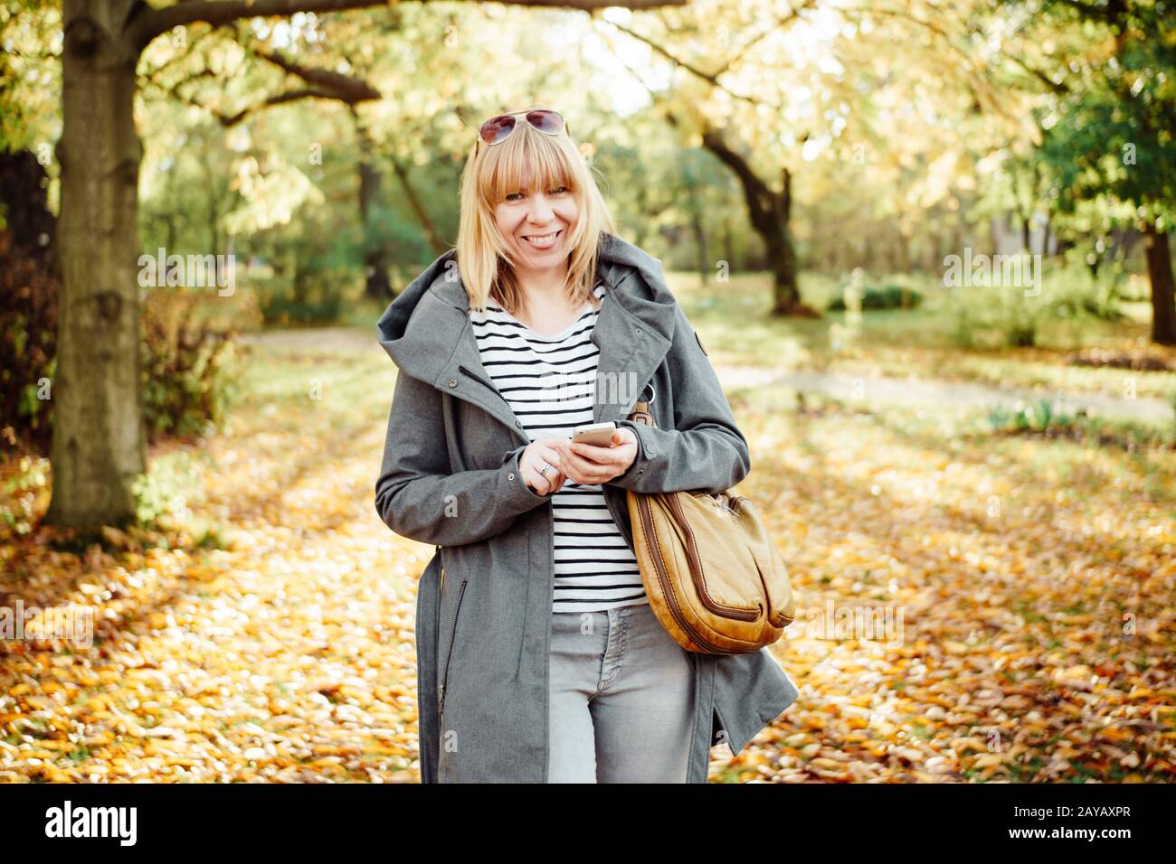 Happy blonde femme dans une forêt d'automne ou le parc des sms avec son téléphone mobile. La communication, de la technologie et de l'extérieur concept. Banque D'Images