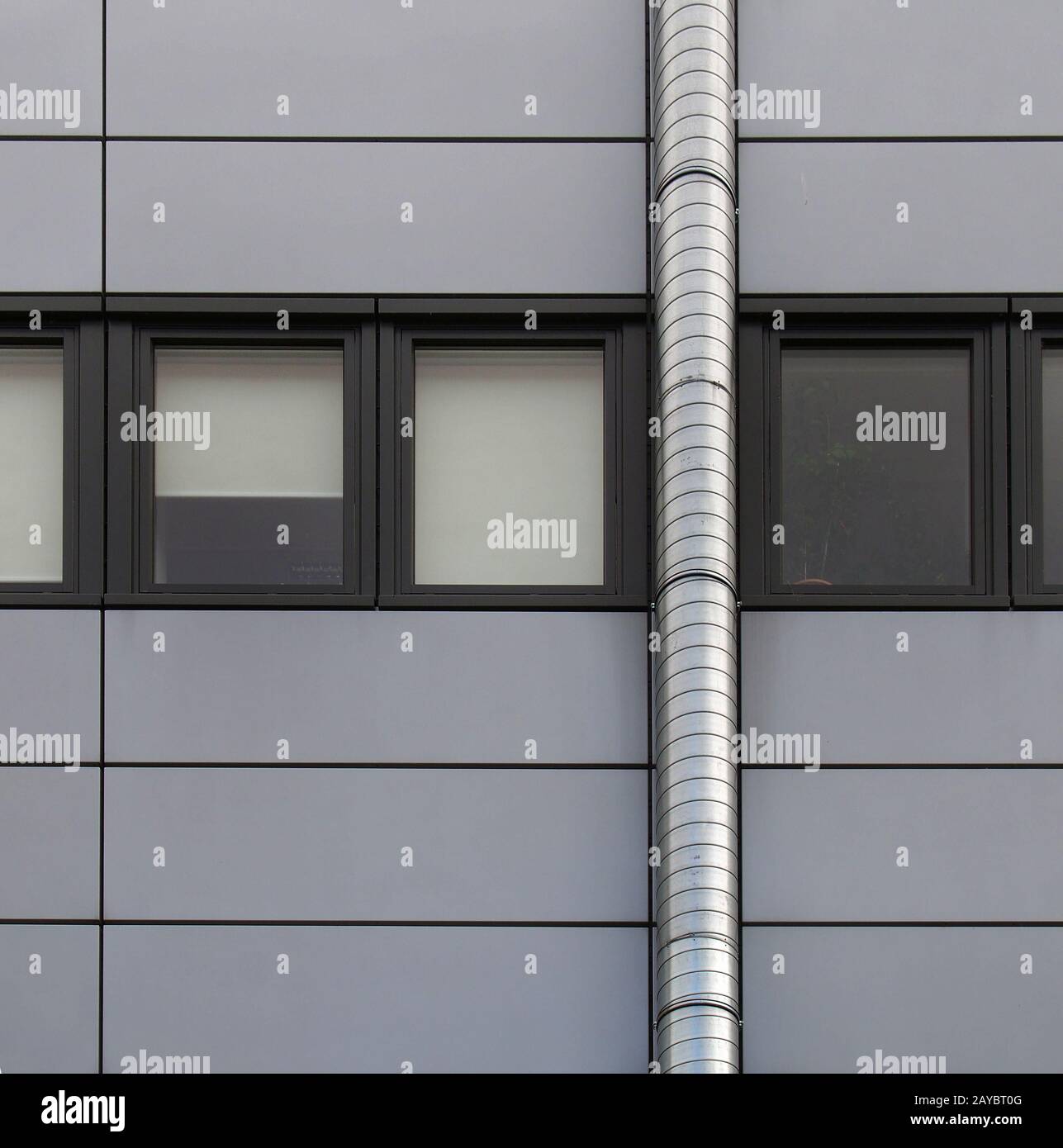 panneaux gris de revêtement sur un bâtiment industriel avec une rangée de cadres de fenêtre arrière et un tuyau en argent qui s'enorne le mur Banque D'Images