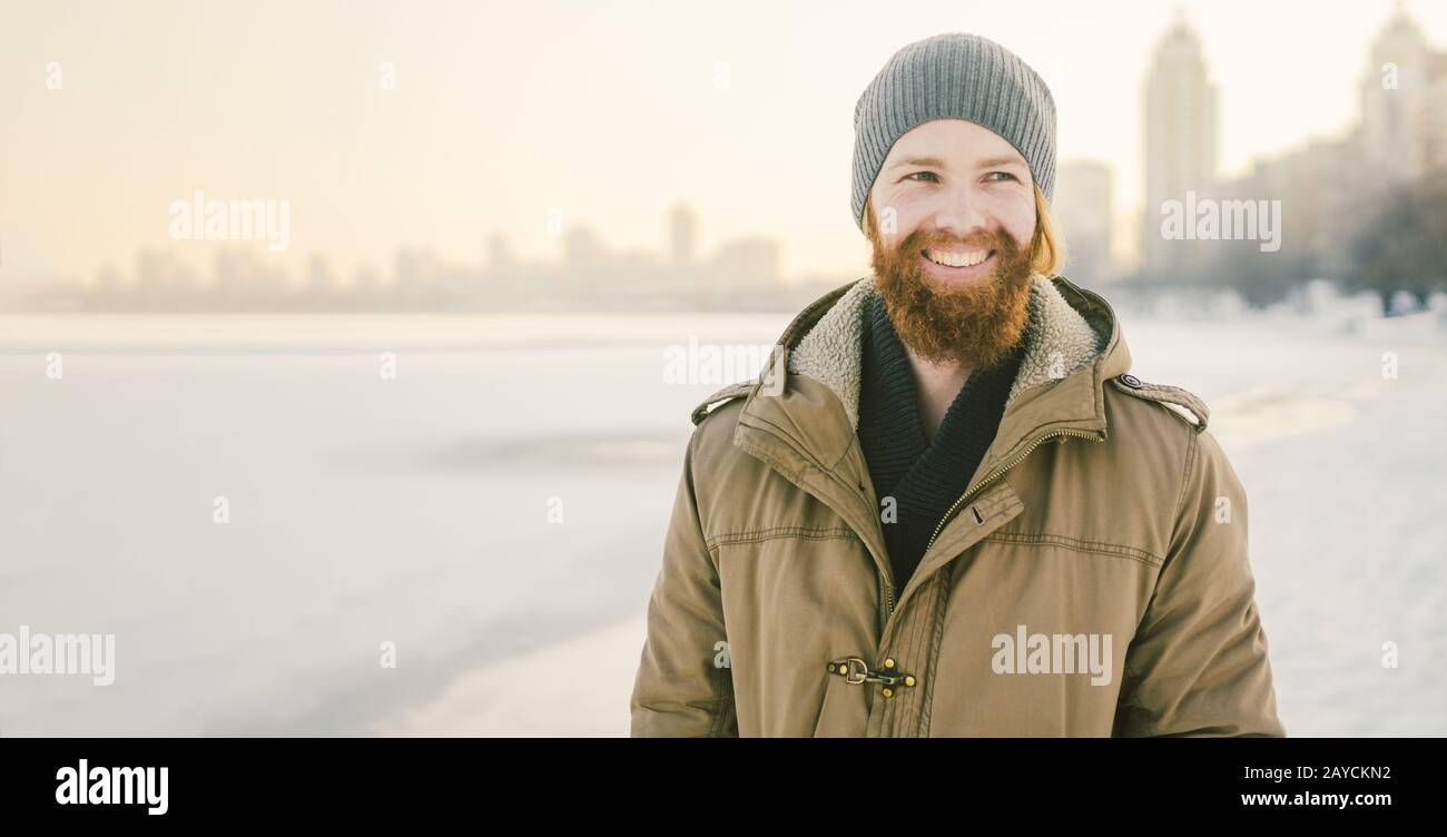 Gros plan caucasien jeunes cheveux rouges et barbe mâles dans un chapeau et un manteau de parc posant le modèle d'hiver sur un fond d'un lac à Banque D'Images