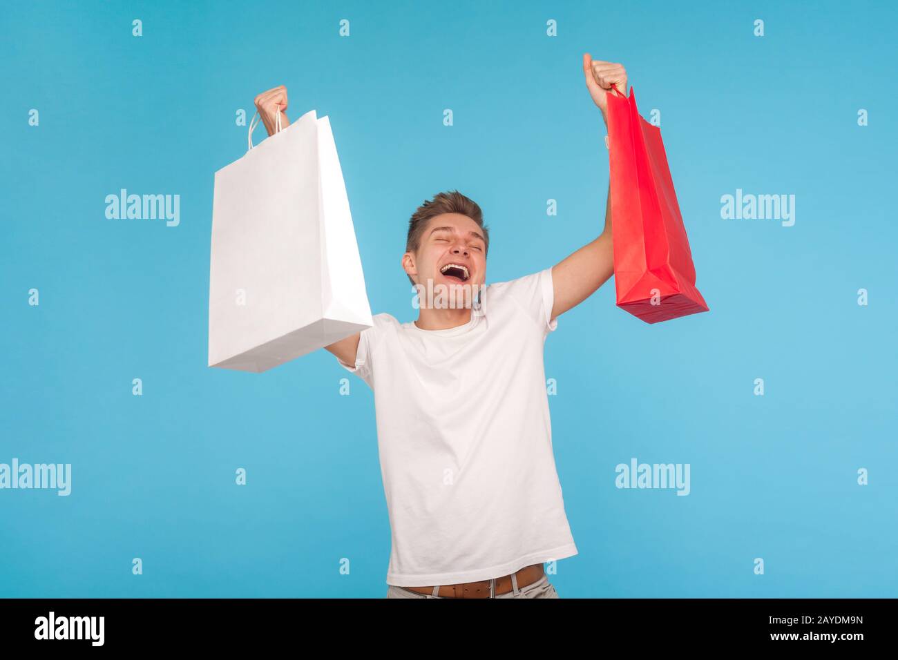 Un homme écostatique surjoé dans un t-shirt criant avec enthousiasme et soulevant des paquets, tenant des sacs de shopping, satisfait de l'achat et favorable pays Banque D'Images