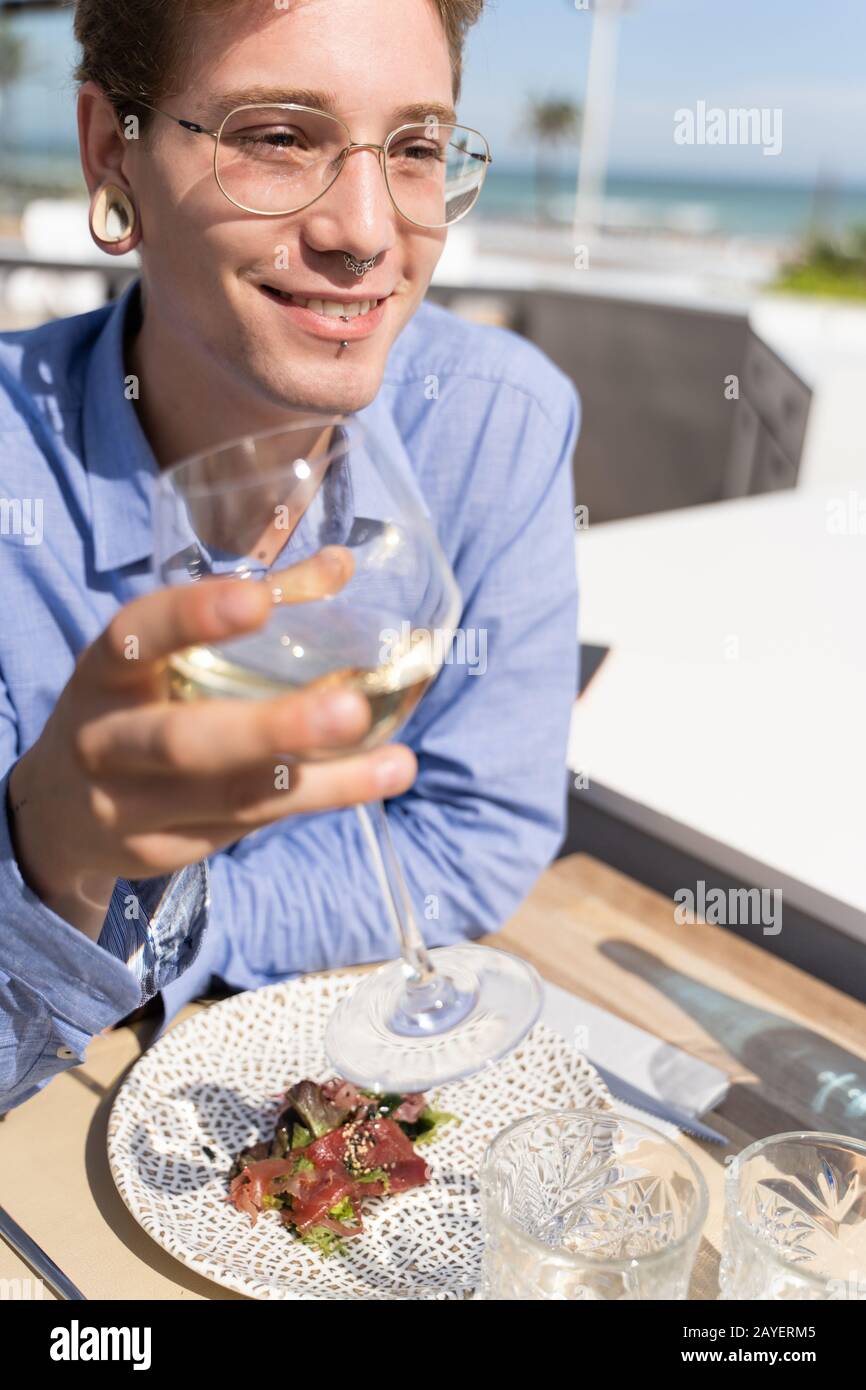 Photo verticale d'un jeune homme avec percées et verres avec un verre de vin blanc dans sa main et une assiette avec salade et saumon avec la table d'un Banque D'Images