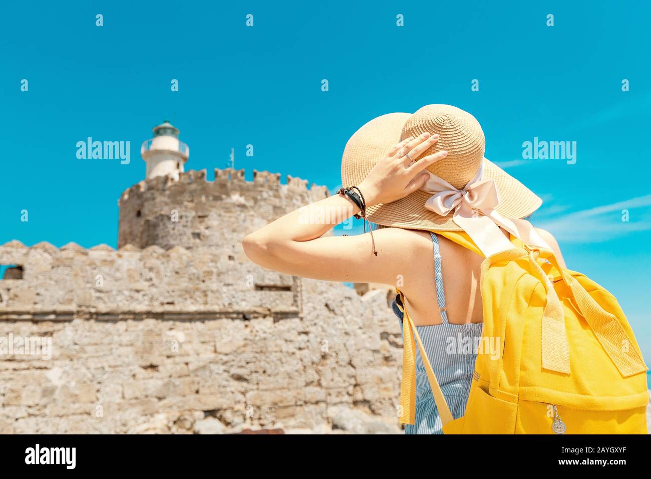 Heureuse femme de tourisme en vacances posant avec chapeau et sac à dos  devant le vieux fort ruiné et phare à Rhodes, Grèce Photo Stock - Alamy