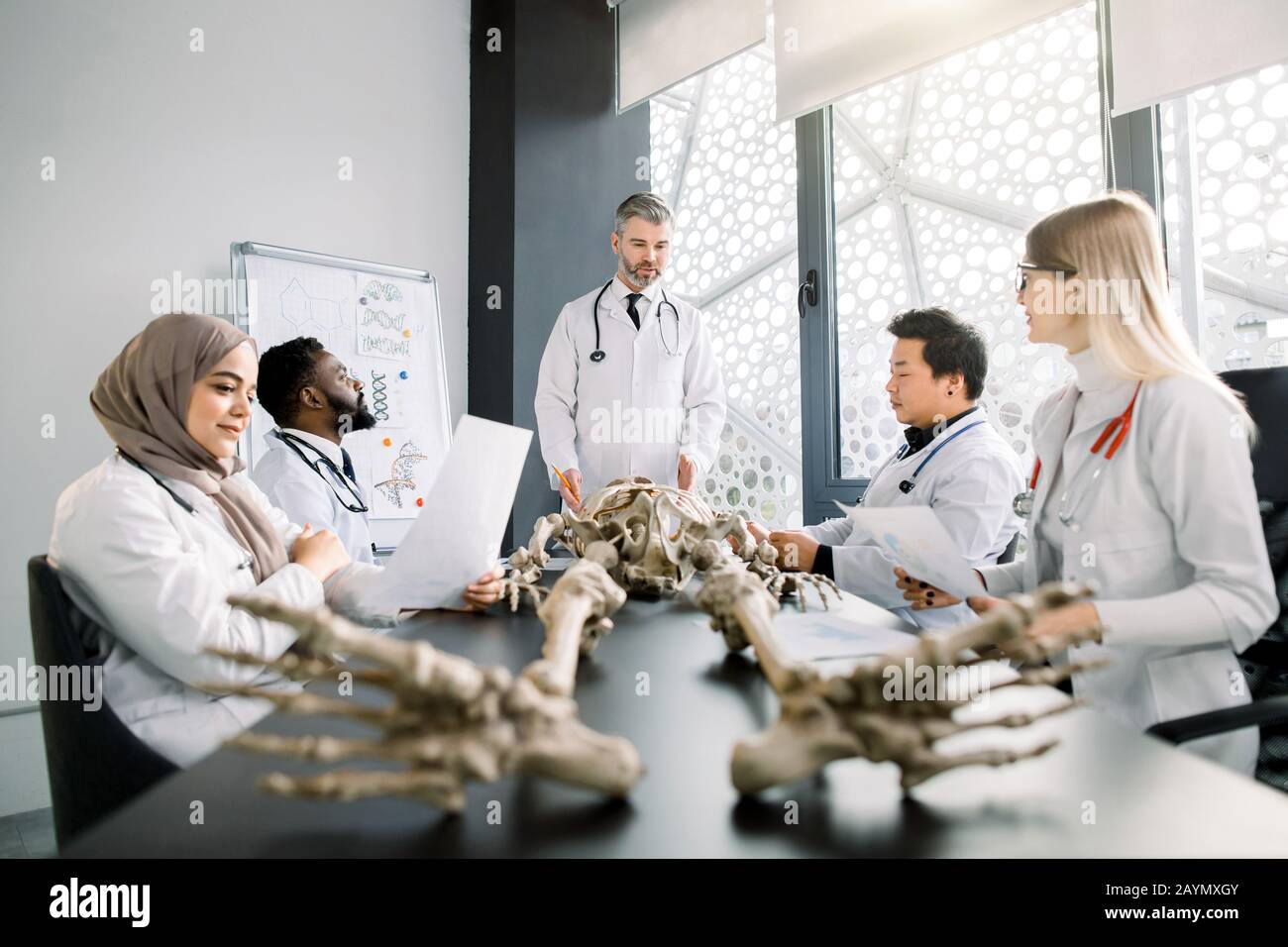 Beau médecin d'âge moyen, professeur d'anatomie, expliquant l'anatomie des os en utilisant le modèle de squelette pour divers élèves en couches blanches. Médecine, éducation Banque D'Images