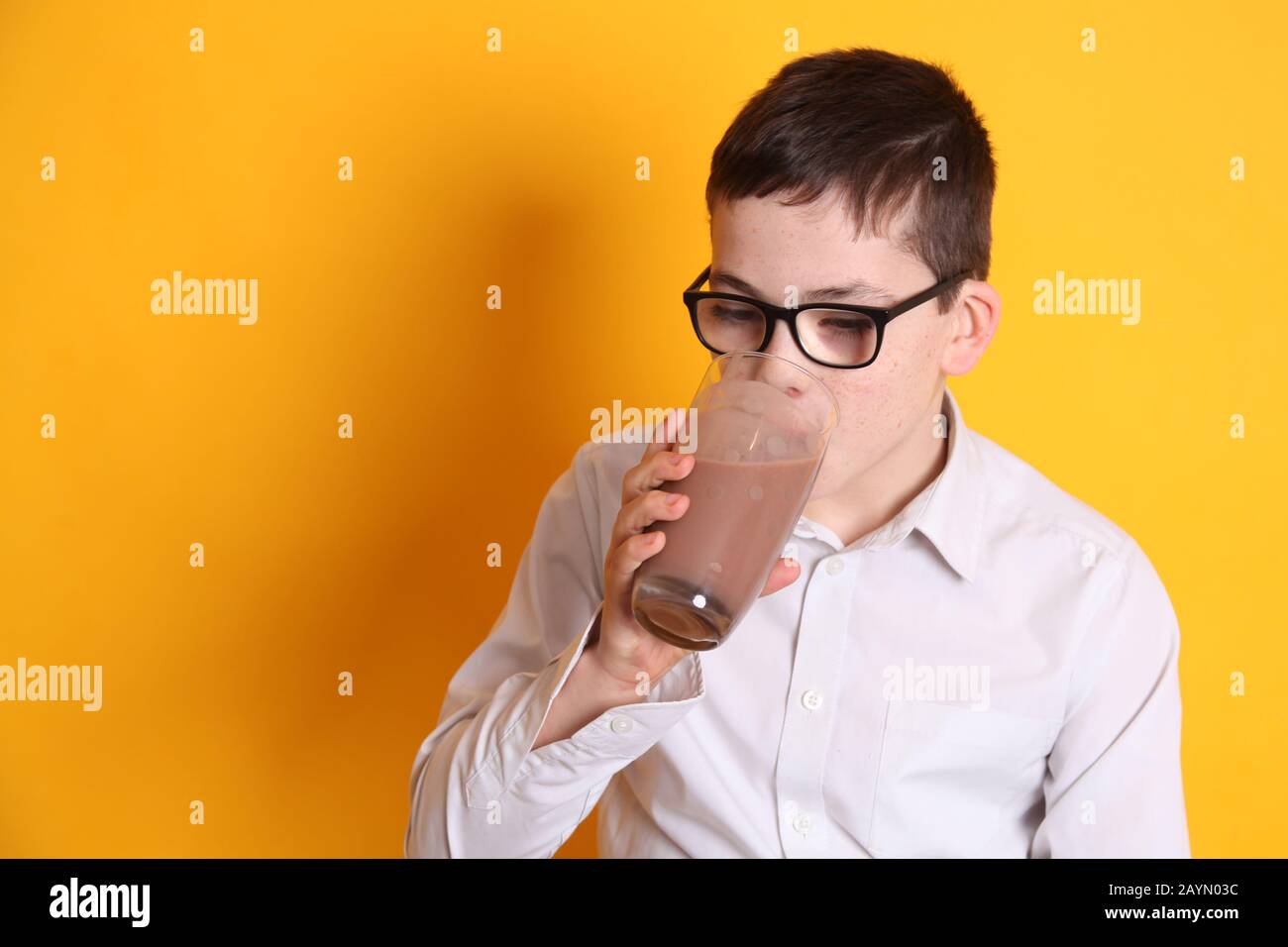 Un jeune garçon de 8 ans boit un verre de lait au chocolat avec une lèvre supérieure recouverte de chocolat sur fond jaune Banque D'Images