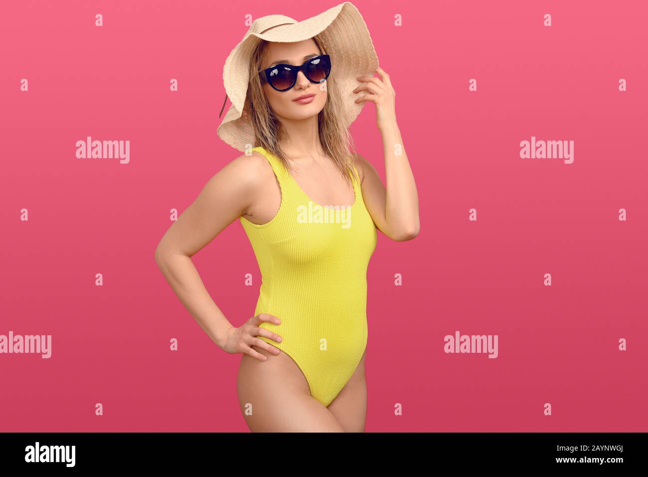 Jeune femme blonde sexy et tendance en maillot de bain jaune, chapeau de soleil à paille de disquette et lunettes de soleil se posant sur un fond de studio rose avec la main sur la hanche dans une thre Banque D'Images