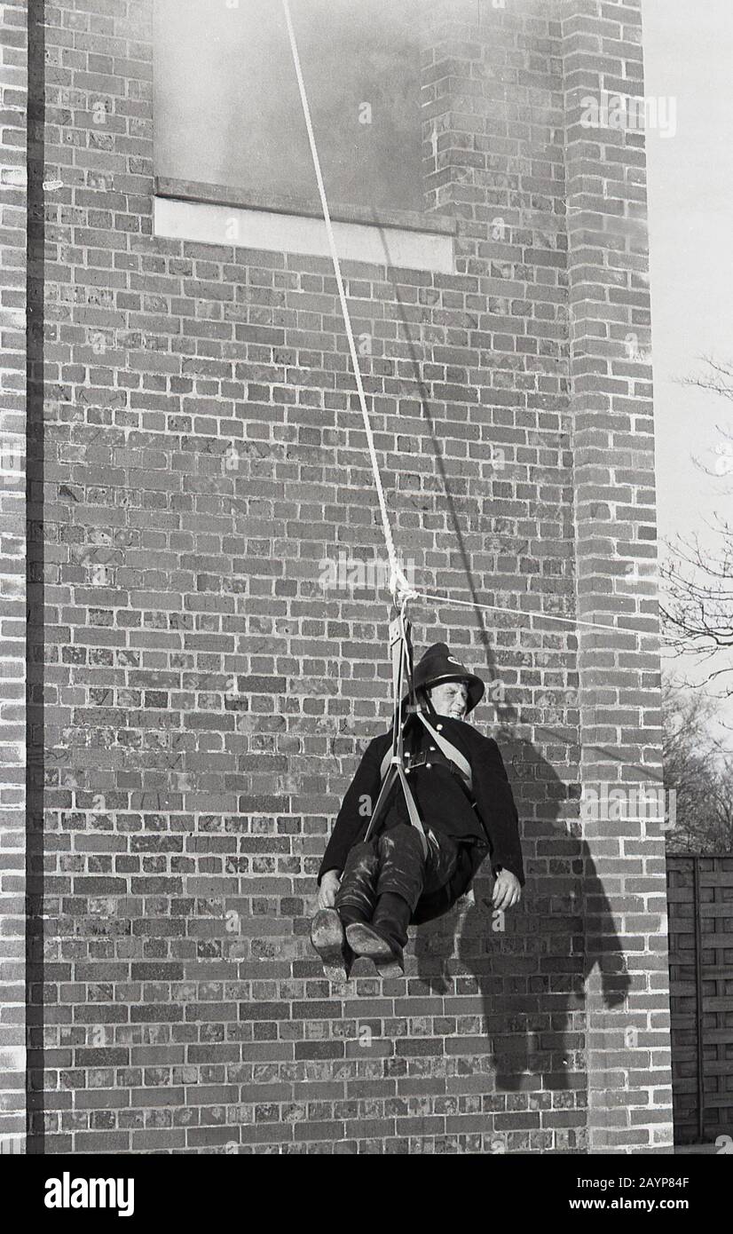 Années 1960, historique, pompier en uniforme et casque de l'époque, participant à un exercice d'entraînement dans le parc de la caserne de pompiers, assis dans un palan, une poulie et une corde, étant évacué d'une tour de forage fumante, Fleetwood, Angleterre, Royaume-Uni. Banque D'Images