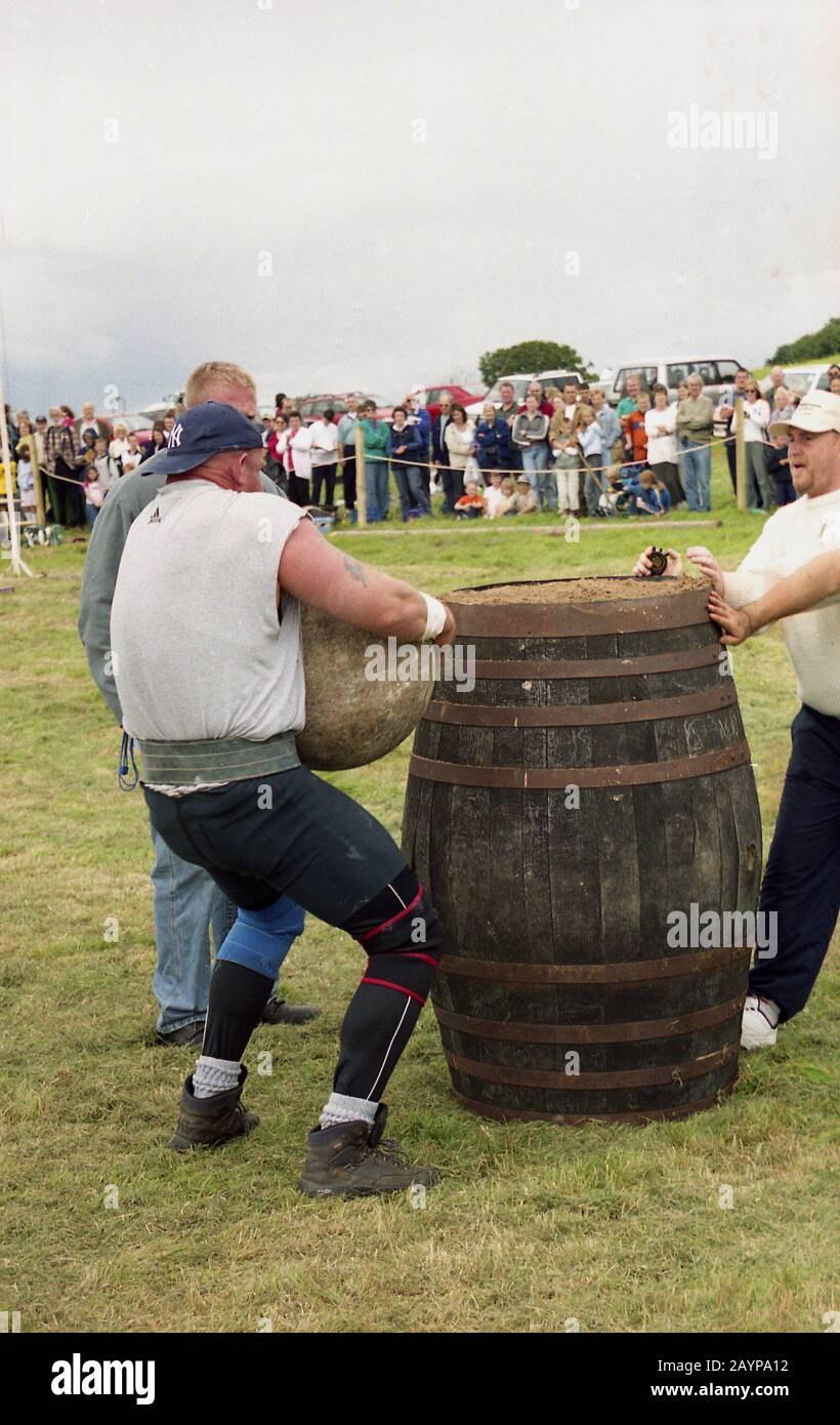 1980, « compétition masculine la plus féroce »... en dehors d'un champ d'herbe surveillé par des spectateurs, un concurrent masculin soulevant une grosse boule de pierre ou une sphère du sol dans le but de la soulever sur le sommet d'un vieux fût de whisky en bois dans un concours d'homme le plus fort, Highlands, Écosse, Royaume-Uni. L'événement peut être évalué sur le poids de la pierre et le temps pris ou les deux. Banque D'Images