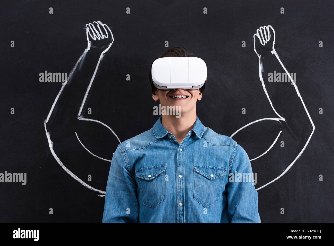 heureux jeune homme utilisant le casque de réalité virtuelle, avec des bras musculaires s'appuyant sur le tableau noir Banque D'Images