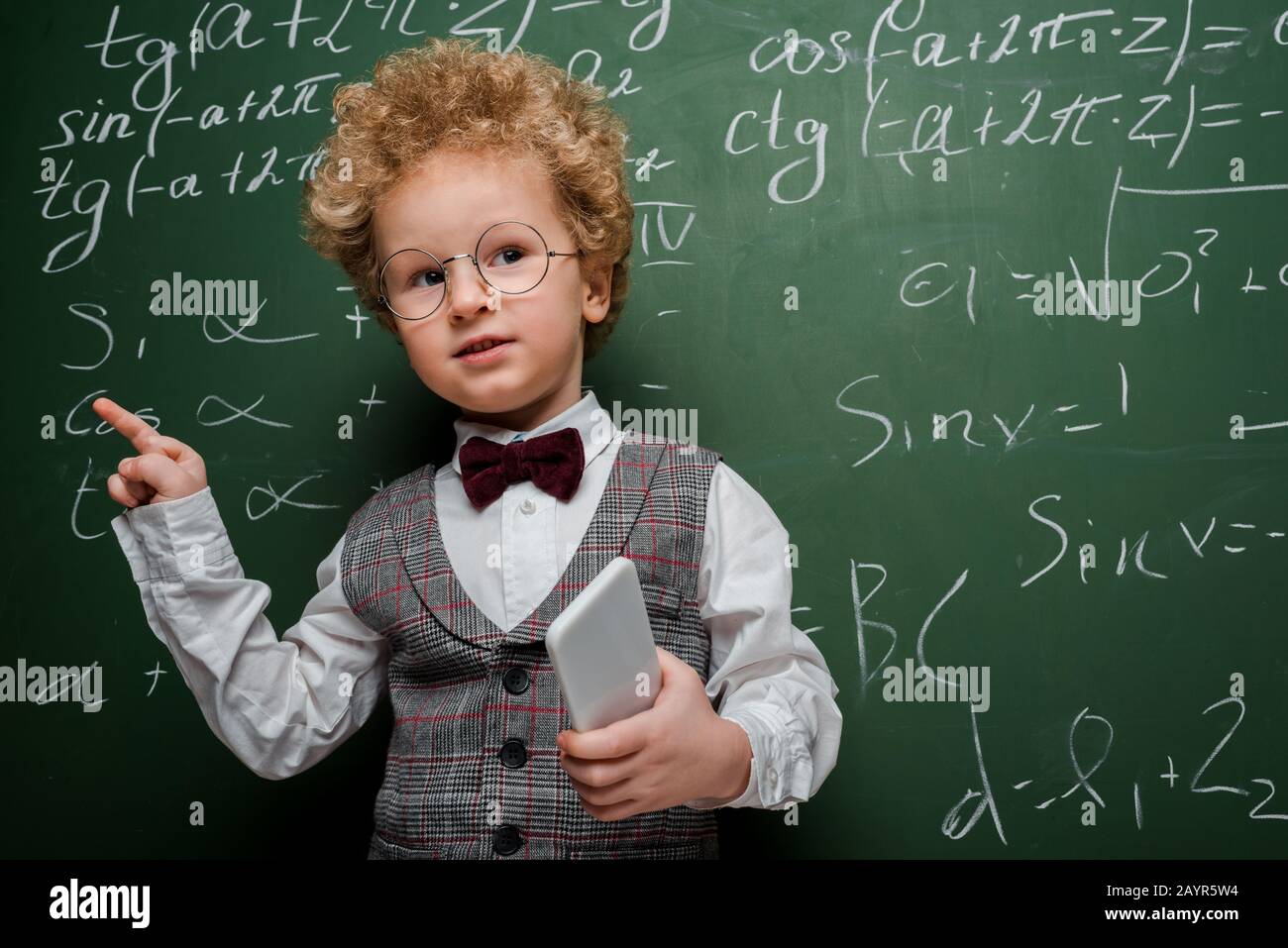 enfant intelligent en costume et noeud papillon tenant le smartphone et pointant avec le doigt près du tableau de surveillance avec des formules mathématiques Banque D'Images