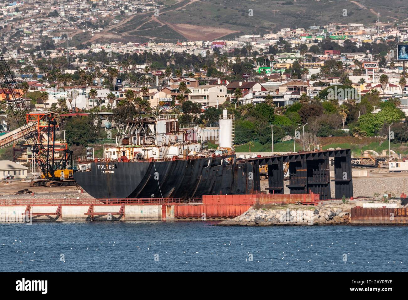 Ensenada, Mexique - 17 janvier 2012: Barge noire Toltec dans un quai sec dans le port. Soutenu par le paysage urbain blanc sur la pente de la montagne. Bleu baie d'eau à l'avant. Banque D'Images