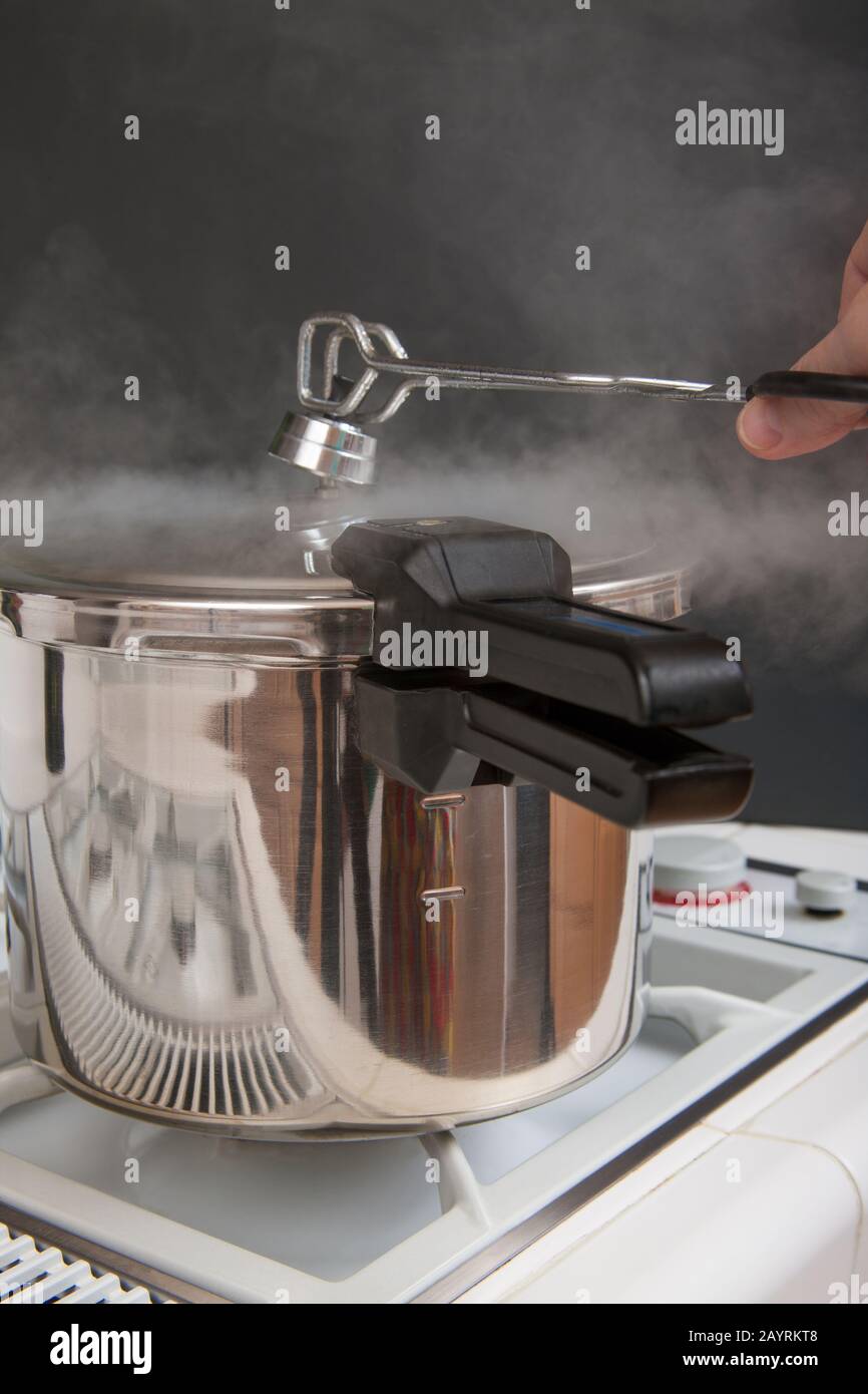 Une cuisinière à pression sur une cuisinière avec vapeur s'échappant lorsqu'une femme utilise des pinces pour retirer le dessus et laisser la vapeur s'échapper Banque D'Images