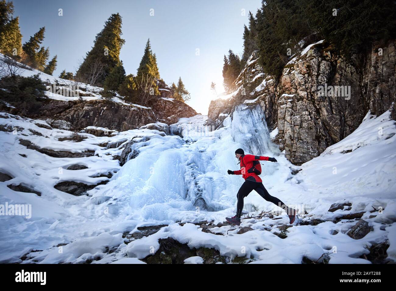 Ancien coureur avec barbe grise et veste rouge sautant près de la cascade gelée aux montagnes en hiver. Sky Running et Trail Running activité extérieure co Banque D'Images