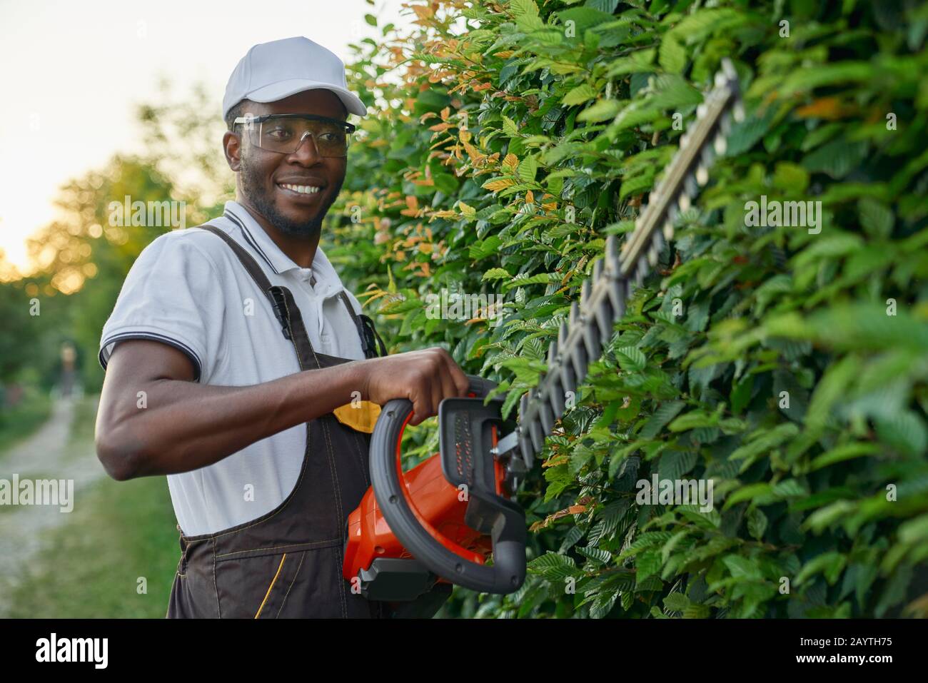 Un jardinier africain souriant portant une combinaison spéciale avec des lunettes de protection travaillant avec des équipements de jardin professionnels à l'extérieur. Jeune homme utilisant un taille-haie au travail Banque D'Images