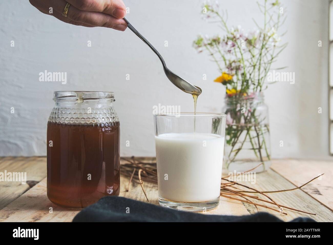 La main de la femme verse du miel naturel sur un verre de lait sur une table en bois dans une cuisine rustique contre la décoration florale. Espace de copie vide. Banque D'Images