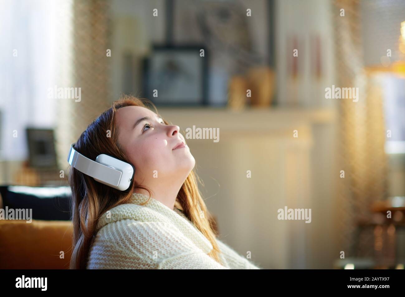 jeune femme moderne avec cheveux rouges dans un pull blanc écoutant la musique avec un casque et regardant sur l'espace de copie tout en étant assis près du canapé en mode Banque D'Images