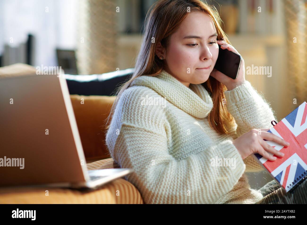 Inquiète jeune femme moderne avec des cheveux rouges dans un pull blanc avec des couleurs d'ordinateur portable et de portable du drapeau britannique parlant sur un téléphone portable dans le hous moderne Banque D'Images