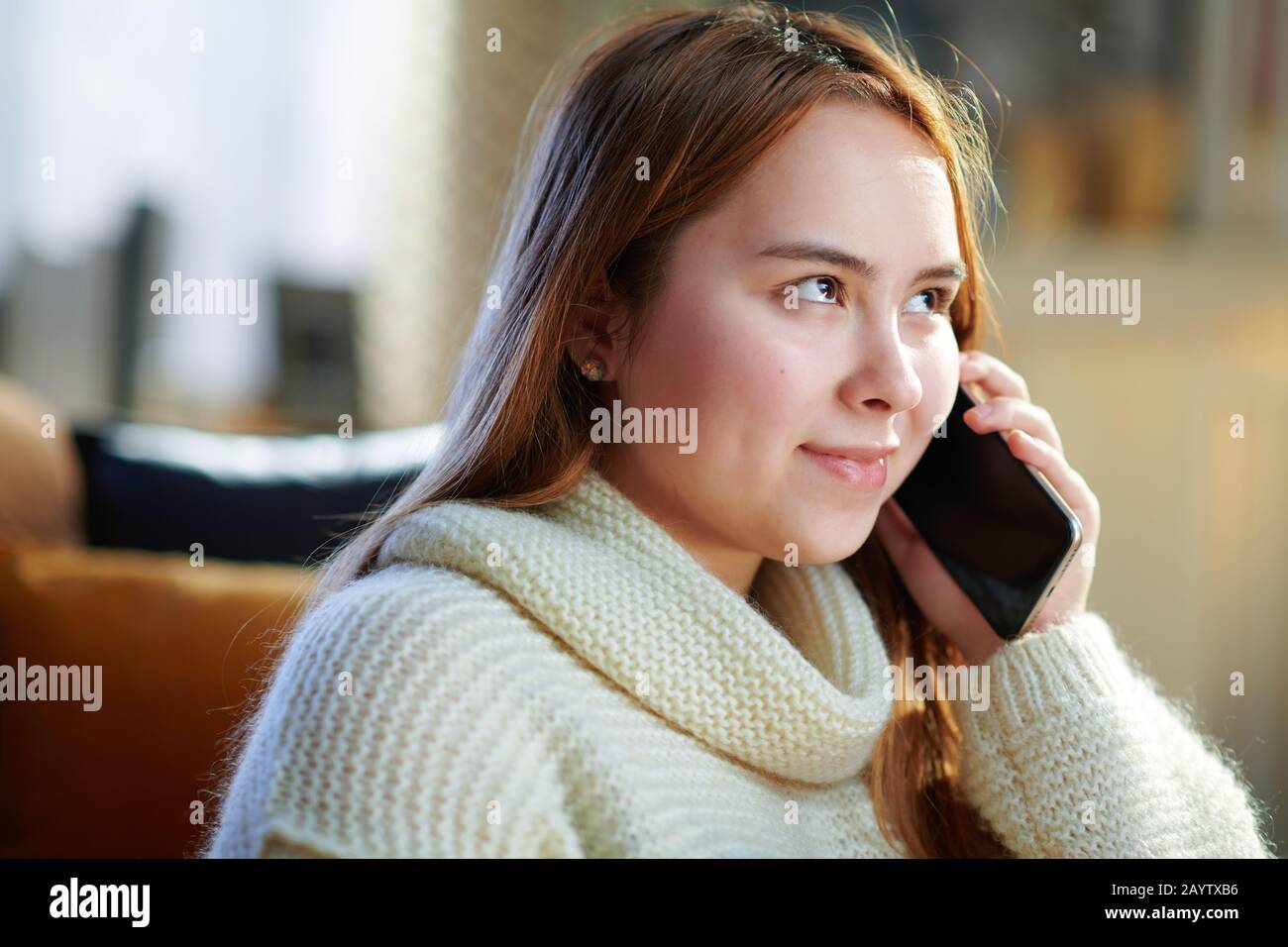 heureuse jeune femme moderne avec des cheveux rouges dans un pull blanc dans la maison moderne en hiver ensoleillé parler sur un téléphone cellulaire. Banque D'Images