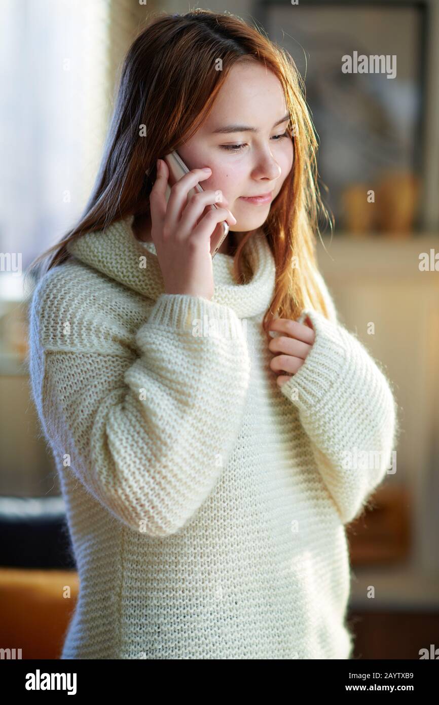 jeune femme moderne avec des cheveux rouges dans un pull blanc dans la maison moderne en hiver ensoleillé à l'aide d'un téléphone cellulaire. Banque D'Images