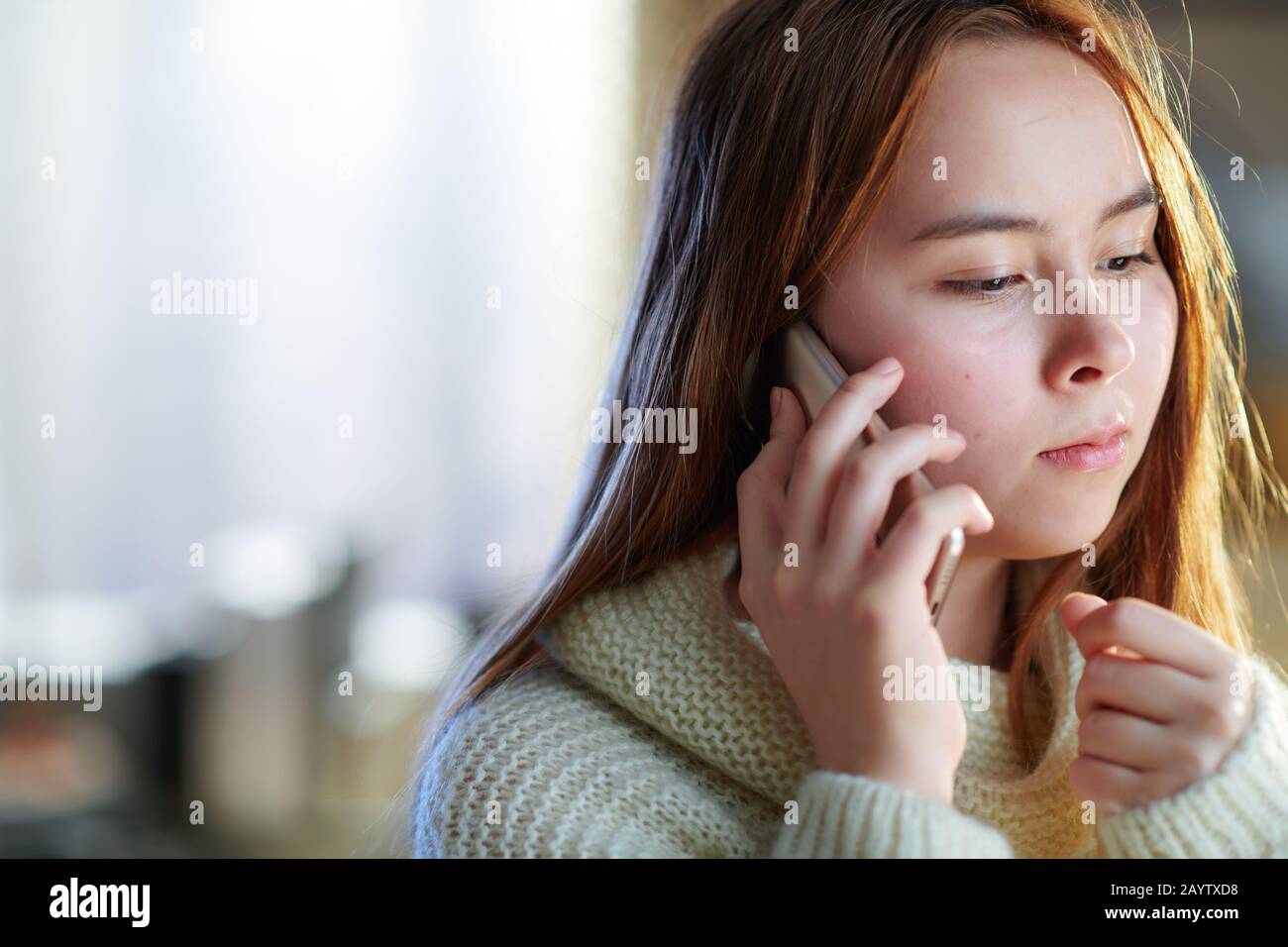jeune femme moderne avec cheveux rouges dans un pull blanc dans le salon moderne en hiver ensoleillé parlant sur un téléphone mobile. Banque D'Images