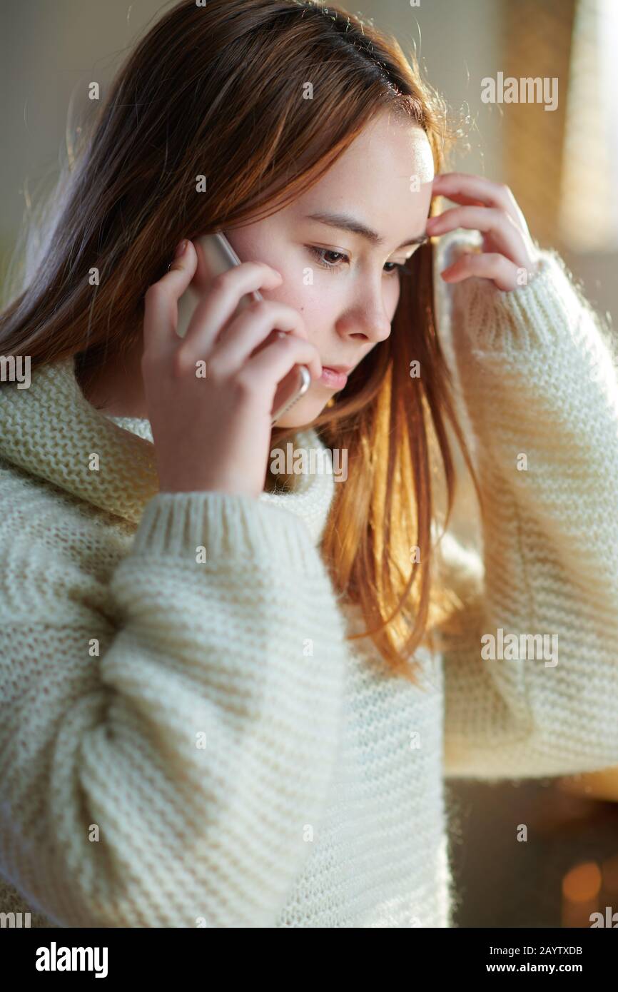 une jeune fille moderne avec des cheveux rouges dans un pull blanc dans la maison moderne en hiver ensoleillé parlant sur un téléphone cellulaire. Banque D'Images