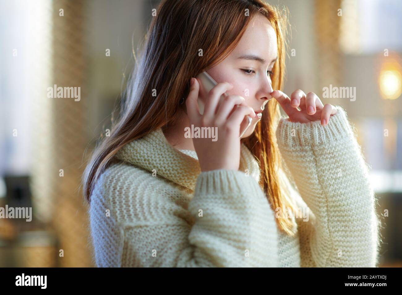 une adolescente moderne avec des cheveux rouges dans un pull blanc dans la maison moderne en hiver ensoleillé parlant sur un téléphone cellulaire. Banque D'Images