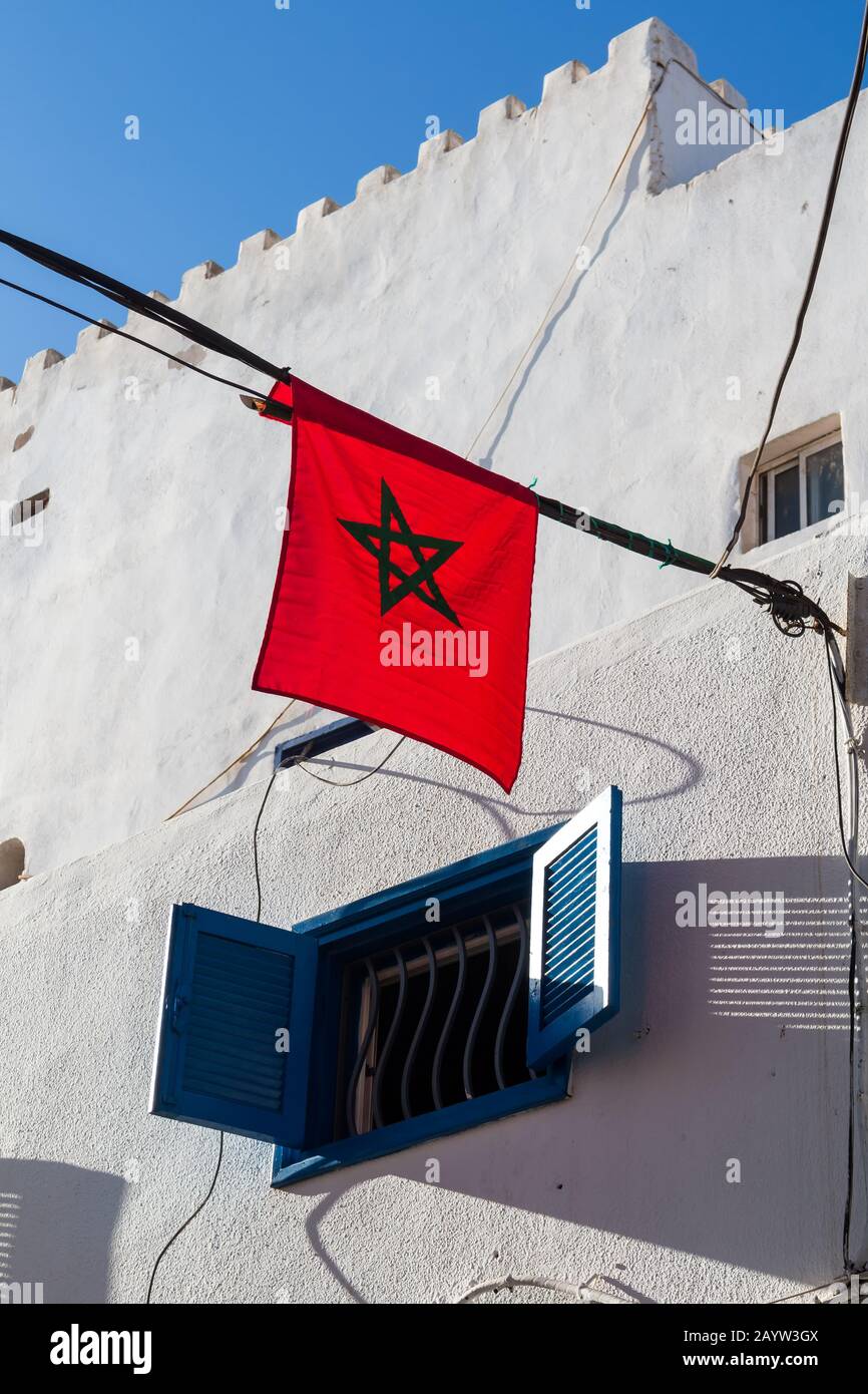 Drapeau marocain à la lumière du soleil, suspendu dans la rue. Façade blanche d'une maison avec fenêtre avec volet bleu ouvert. Ligne du ciel bleu. Essaoui Banque D'Images