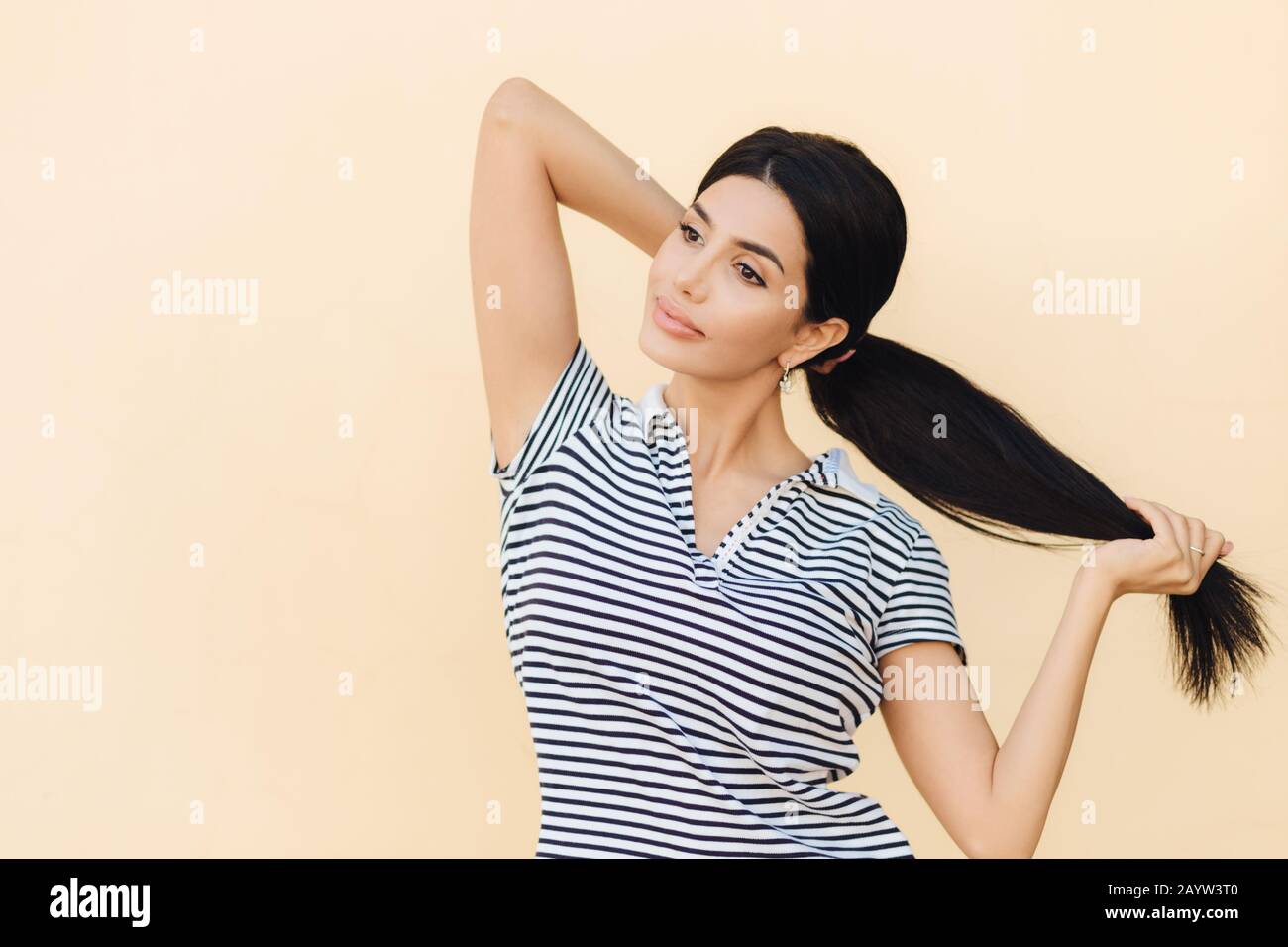 La femelle à poil sombre pensif fait de la queue de poney, a des lèvres pleines et une apparence agréable, porte une tenue décontractée, se tient sur le fond du studio, focalisé sur Banque D'Images