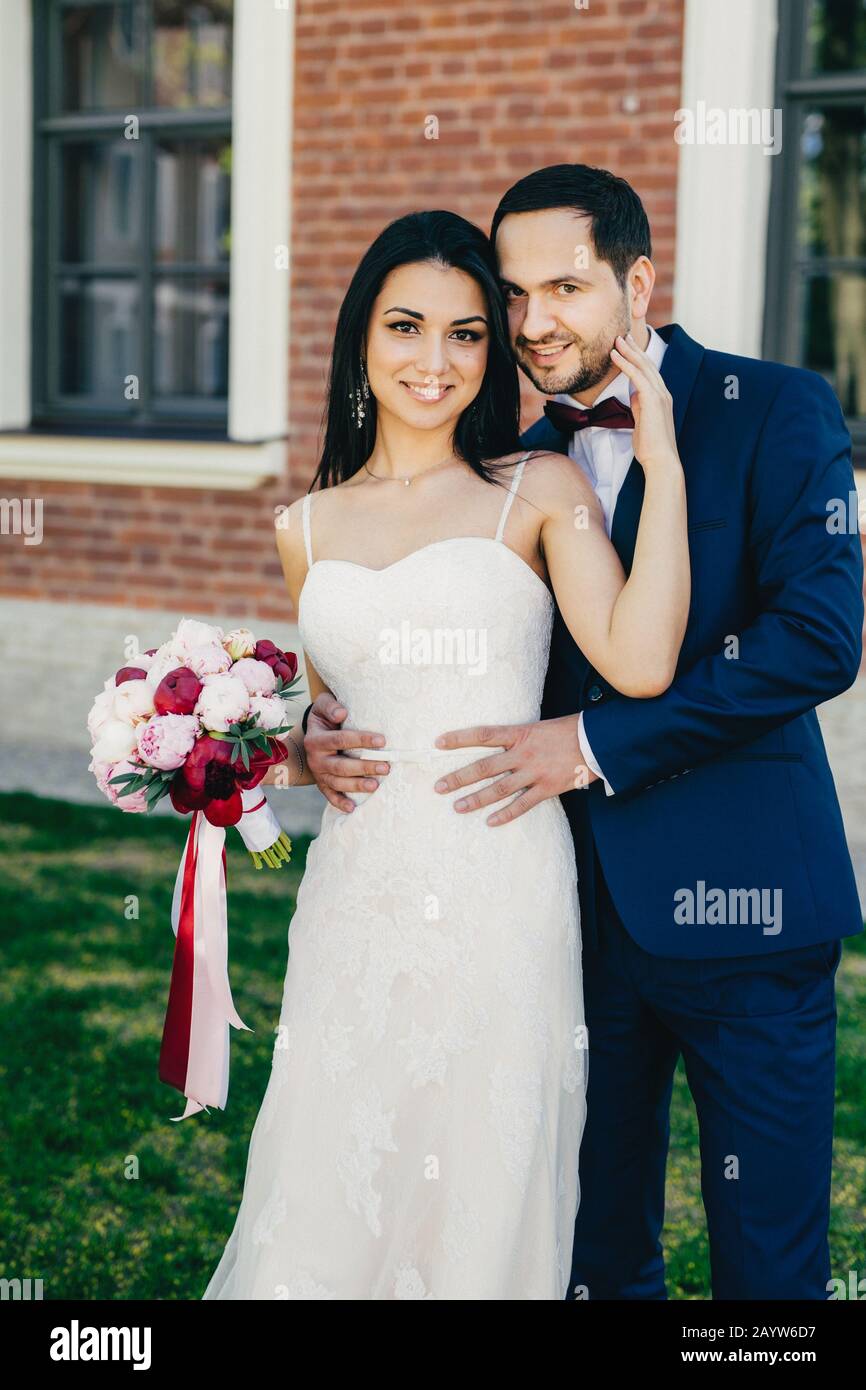 Agreable look jeune femme brunette dans la robe de mariage blanche tient bouquet stands pres de sa belle bridepiece qui porte costume formel stand a l exterieur Photo Stock Alamy