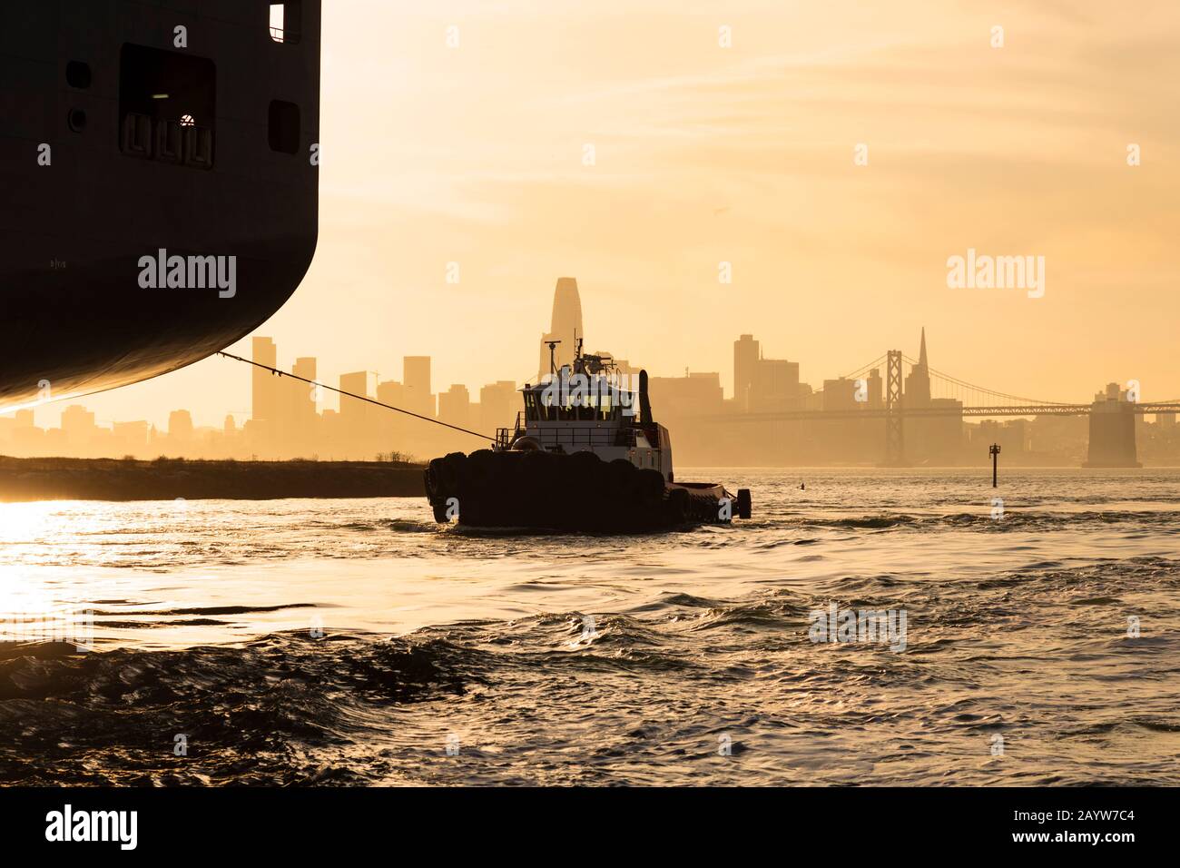Le bateau remorqueur et le bateau à conteneurs se déplacent dans le port d'Oakland contre le soleil couchant et les gratte-ciel de San Francisco. Banque D'Images