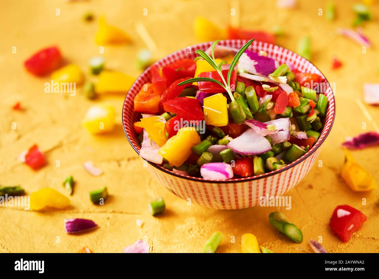 gros plan d'un bol avec un mélange de différents légumes crus hachés, tels que les asperges, l'oignon ou le poivron jaune et rouge, sur une surface dorée texturée Banque D'Images