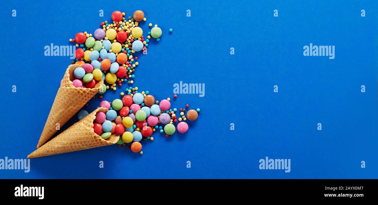 Panorama festif avec bonbons au chocolat enrobés de sucre colorés qui débordent de deux cônes de glace de gaufre sur un fond bleu avec espace de copie Banque D'Images