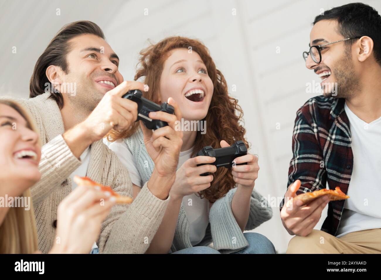 En dessous de la vue de jeune homme excité et de femme jouant au jeu vidéo tandis que leurs amis mangeant la pizza et l'enracinement pour eux Banque D'Images