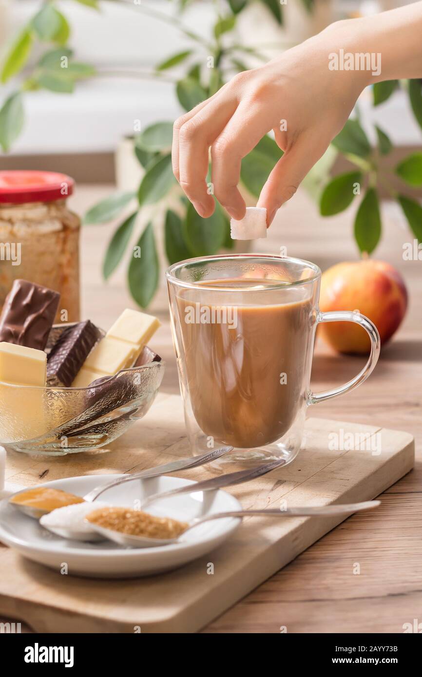 La main du garçon tient un cube de sucre sur un verre de thé ou de café avec du lait. Différents types de bonbons et de sucre se trouvent sur la table. Banque D'Images