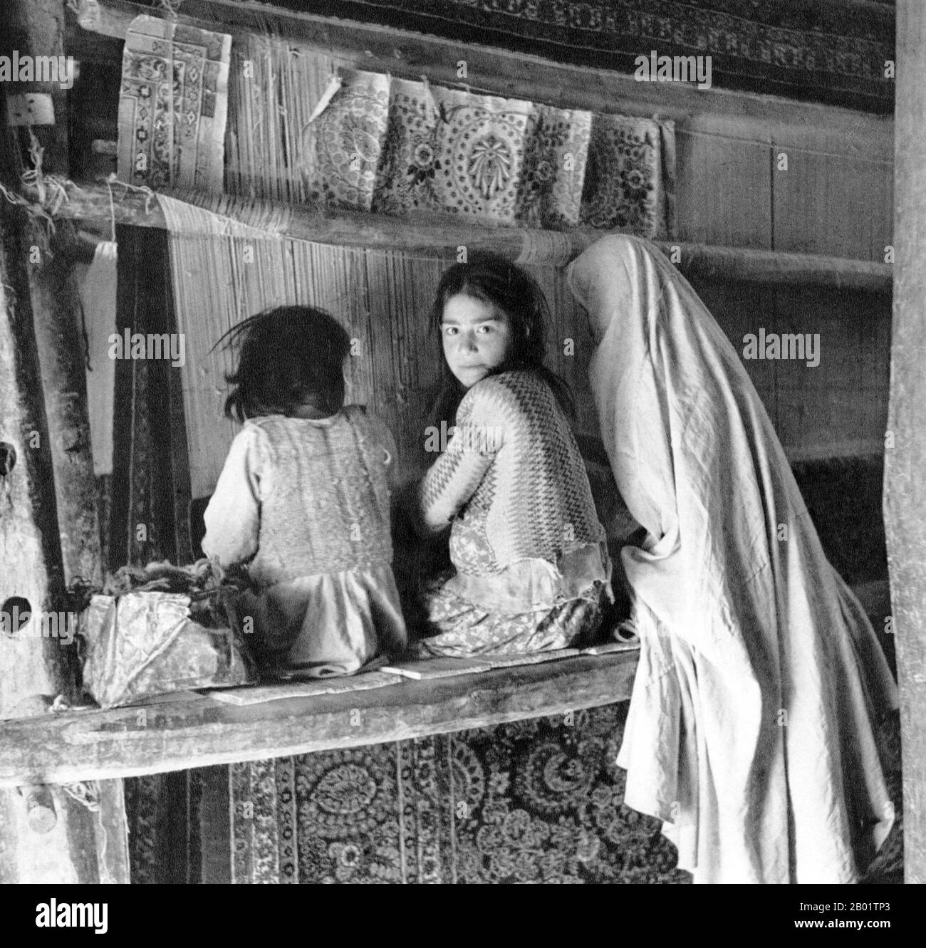 Iran/Perse : femme tissant des tapis avec deux jeunes filles, Hamadan, v. 1920. Hamadān ou Hamedān (vieux persan : Hagmatana, grec ancien : Ecbatana) est la capitale de la province iranienne de Hamadan. Au recensement de 2006, sa population était de 473 149 habitants répartis dans 127 812 familles. Hamedan est considéré comme l'une des plus anciennes villes iraniennes et l'une des plus anciennes du monde. Il est possible qu'elle ait été occupée par les Assyriens en 1100 av. J.-C. ; l'historien grec ancien, Hérodote, déclare qu'elle était la capitale des Mèdes, vers 700 av. J.-C.. Banque D'Images