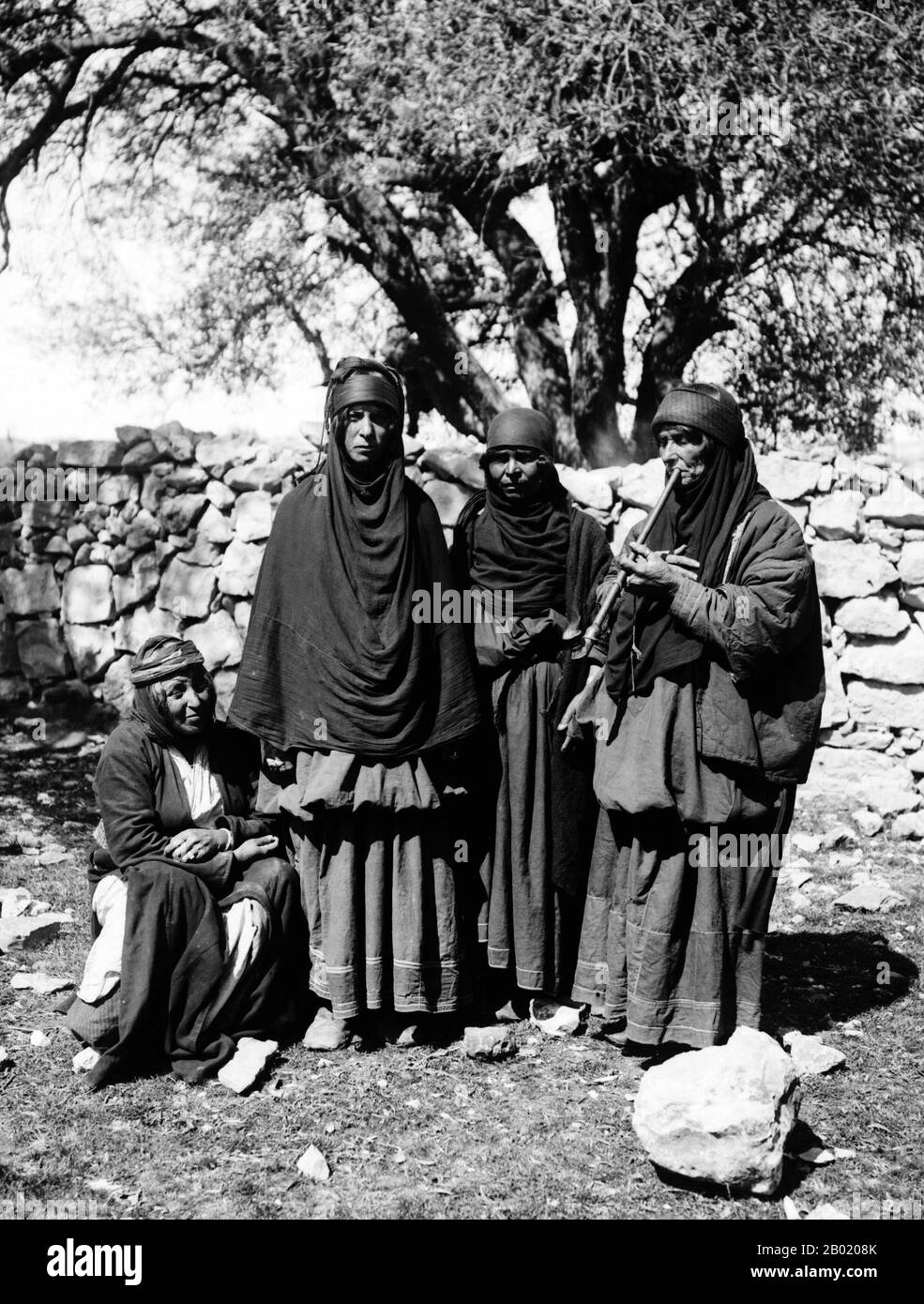 Jordanie : un groupe de bédouines, dont l'une fume une pipe, v. 1900-1920. Les Bédouins font partie d'un groupe ethnique arabe vivant principalement dans le désert, traditionnellement divisé en tribus, ou clans, connu en arabe sous le nom de ʿašāʾir. Le terme bédouin dérive d'une forme plurielle du mot arabe badawī, tel qu'il est prononcé dans les dialectes familiers. Le terme arabe badawī dérive du mot bādiyah, qui signifie désert semi-aride (par opposition à ṣaḥarāʾ qui signifie désert). À partir de la fin du XIXe siècle, de nombreux Bédouins sous domination britannique ont commencé à transiter vers une vie séminomadique. Banque D'Images