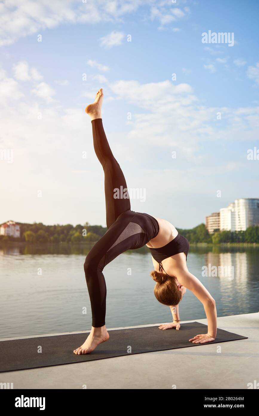 Jeune femme dans des vêtements de sport noirs faisant du yoga ou pilates s'exercer sur tapis près du lac contre l'arrière-plan du paysage urbain sous le ciel bleu. Entraînements le matin, dynamisez votre énergie toute la journée Banque D'Images