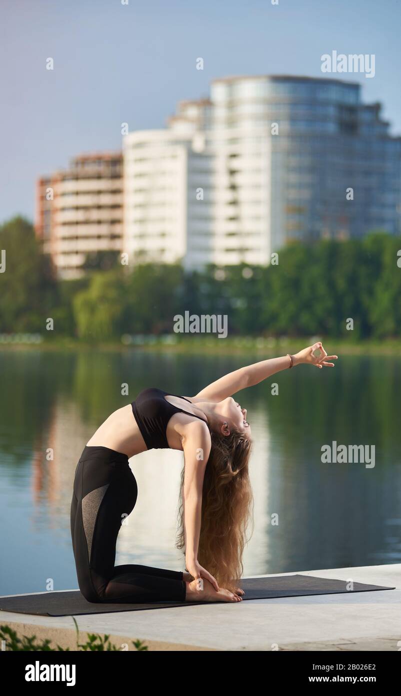 Femme flexible avec cheveux longs pratiquant le yoga à l'extérieur près de la rivière, assis à Camel pose ou Ustrasana. Fille qui travaille dans un pantalon noir, soutien-gorge au tapis contre fond flou des bâtiments de la ville Banque D'Images