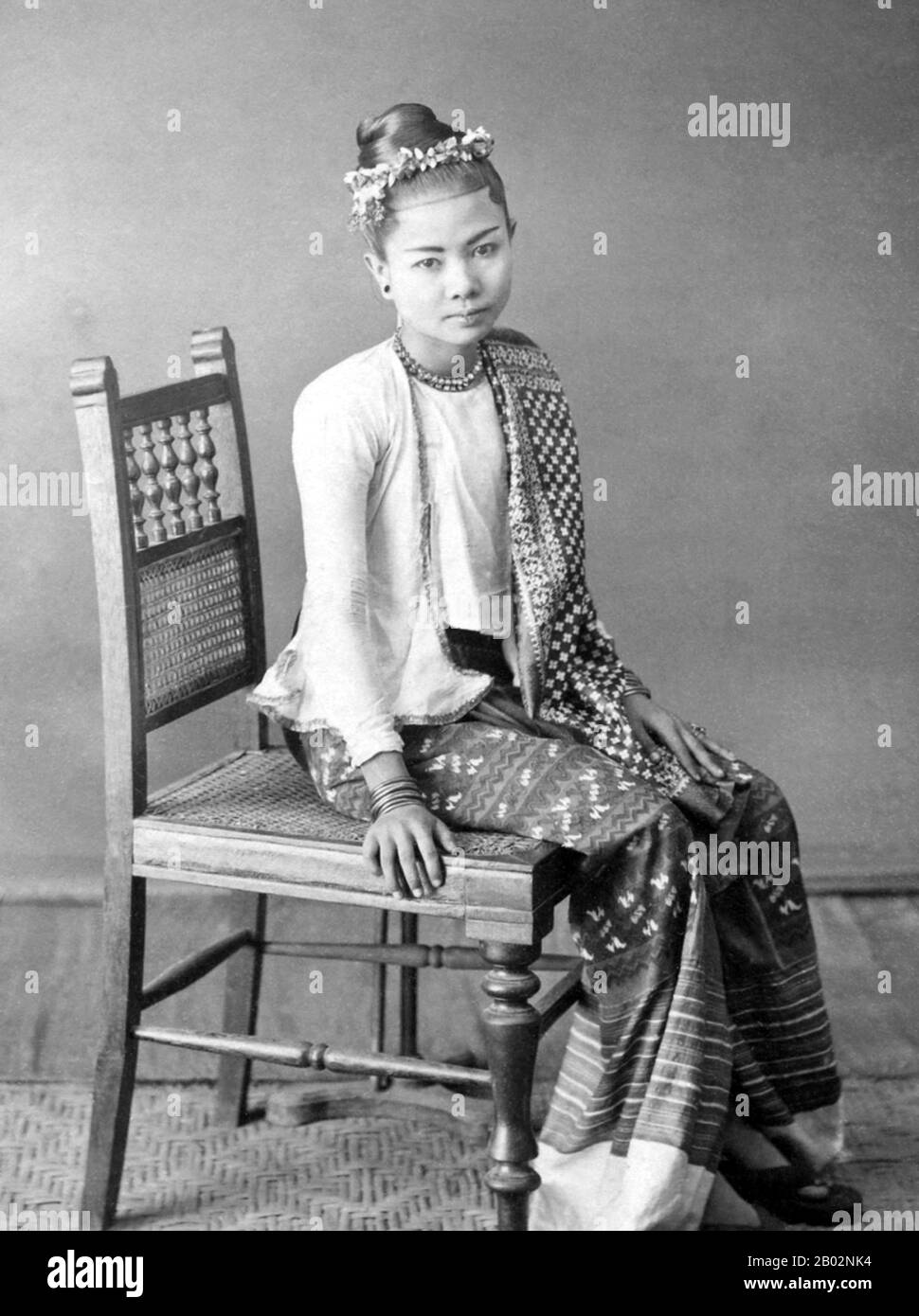 La jeune femme porte une hta-mein rayée (jupe enveloppante) et une veste ajustée de mousseline fine ou de coton connue sous le nom d'éine-gyi. Dans ses cheveux, elle porte un bandeau floral et elle est ornée de colliers, boucles d'oreilles, bracelets et bagues. Pendant la dynastie Konbaung (1752-1885), de riches bijoux, de beaux tissus tels que la soie et des vêtements tels que sa veste étaient réservés aux responsables de la cour et à leurs épouses par la loi. Après la chute de la monarchie birmane en 1885, ils ont été portés par les riches. Cette photo est tirée d'un album qui enregistre des aspects de la tournée de Lord Elgin en Birmanie de novembre à décembre 1898. Banque D'Images