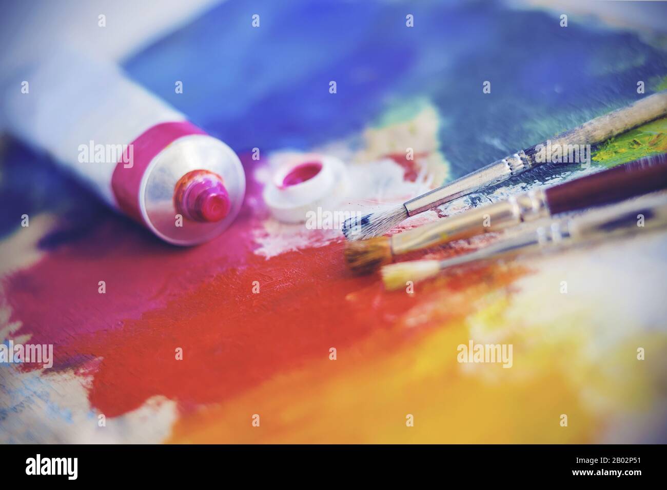 Un tube ouvert de peinture à l'huile rose et trois pinceaux se trouvent sur une palette lumineuse colorée de couleurs différentes. Banque D'Images