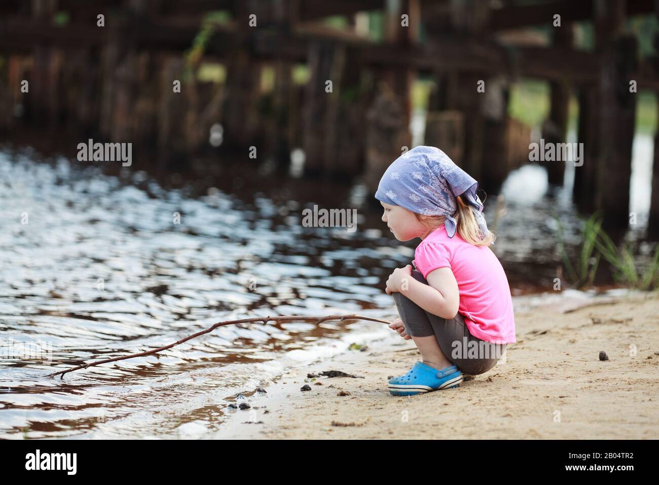Petite fille triste dans un bandana bleu assis sur la rive de la rivière et tire avec une torsion sur l'eau Banque D'Images