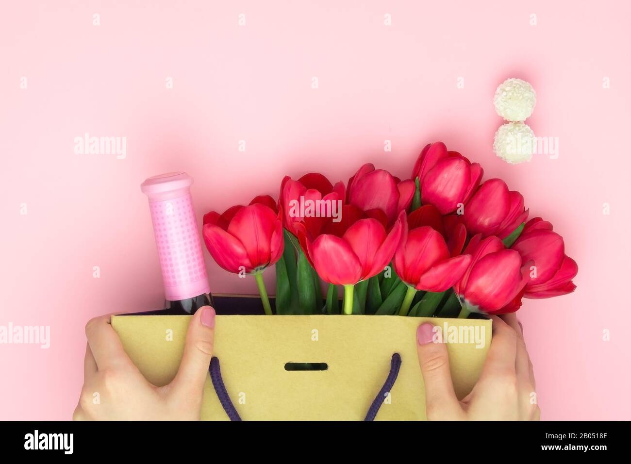 Concept du cadeau avec du vin et des tulipes rouges dans le sac en papier sur le fond rose. Couche plate, vue supérieure, espace de copie. Les mains de la femme tiennent un présent à Banque D'Images