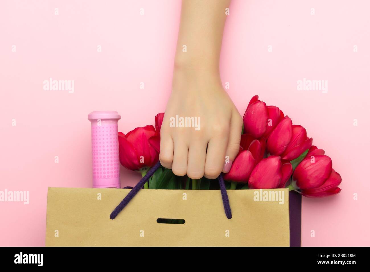 Concept du cadeau avec du vin et des tulipes rouges dans le sac en papier sur le fond rose. Couche plate, vue supérieure, espace de copie. La main de femme est présente à Banque D'Images