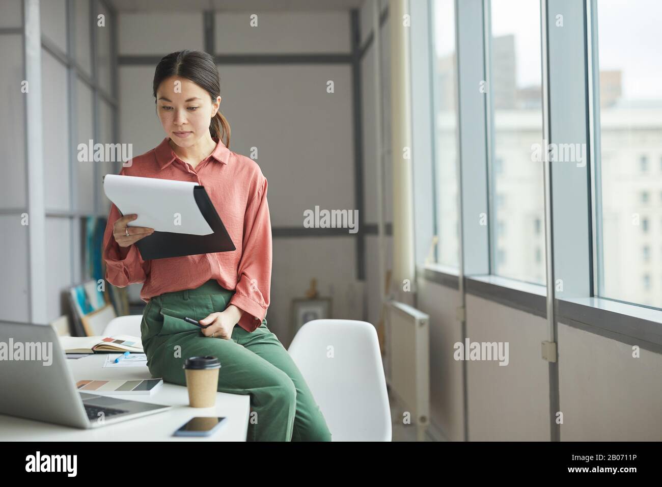 Jeune femme d'affaires assis sur la table avec ordinateur portable et examinant le contrat d'affaires au bureau Banque D'Images