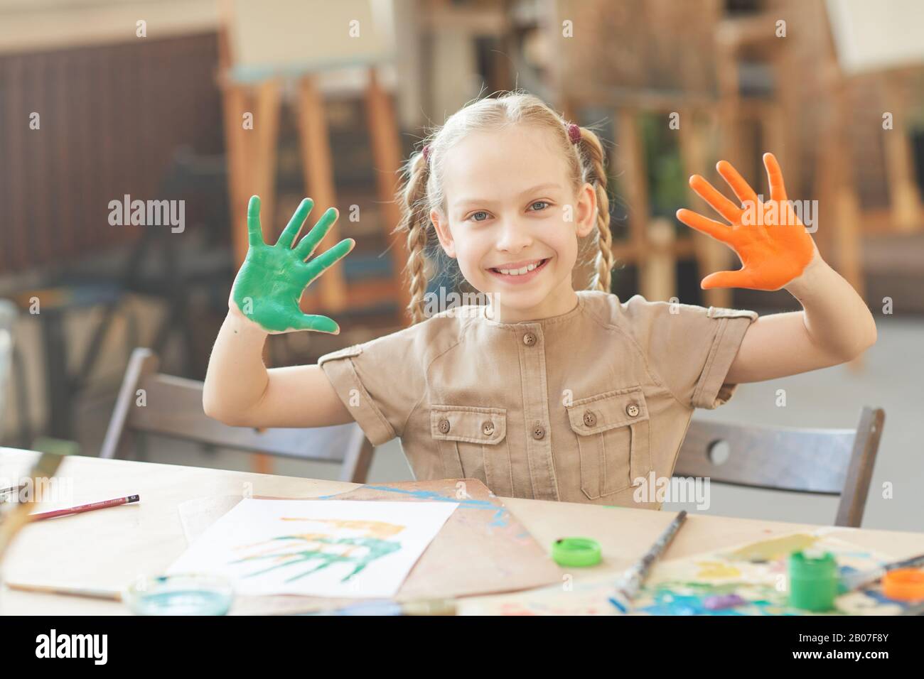 Portrait d'une heureuse fille souriant à l'appareil photo et montrant ses mains dans des peintures tout en étant assis à la table au studio d'art Banque D'Images