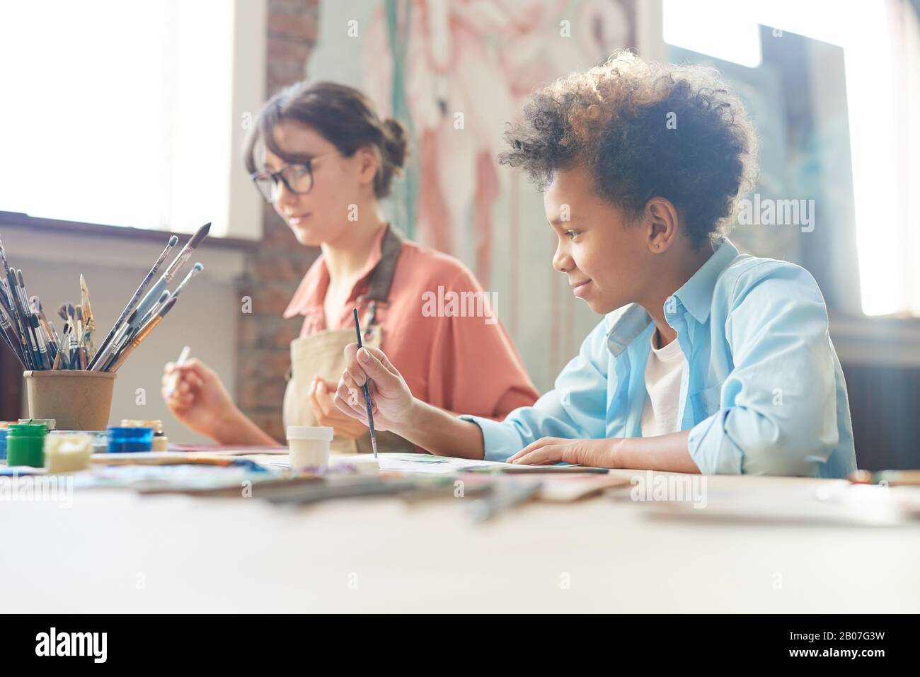 Jeune femme assise à la table avec un garçon africain et ils peignent avec des aquarelles au studio d'art Banque D'Images