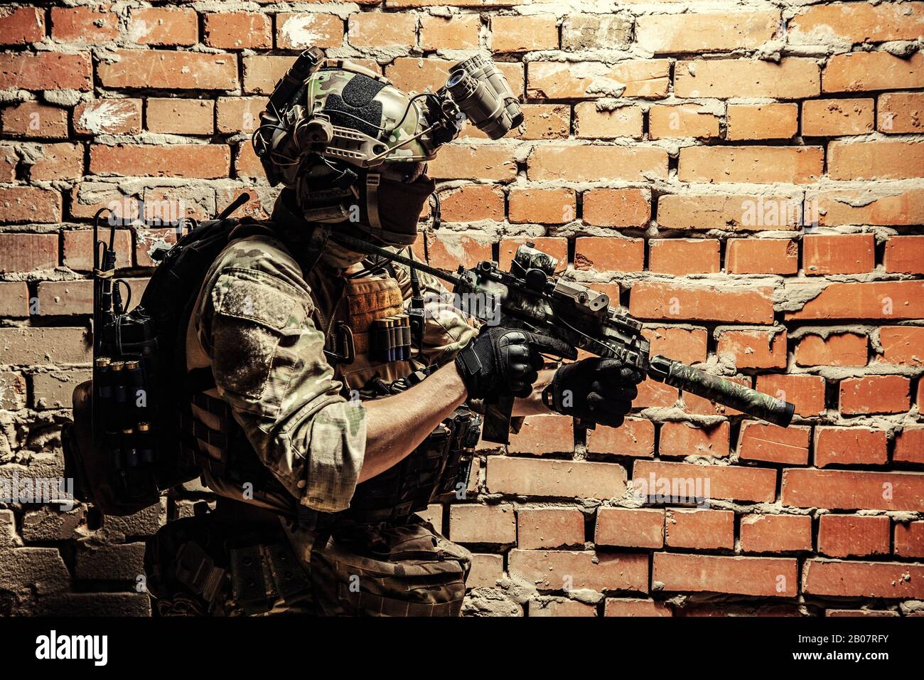 Chasseur d'escouade anti-terroriste, soldat des forces d'élite de l'armée dans des munitions uniformes et tactiques de combat, mini-canon armé, port d'un dispositif de vision nocturne, se déplaçant le long du mur de briques pendant l'opération sqb Banque D'Images