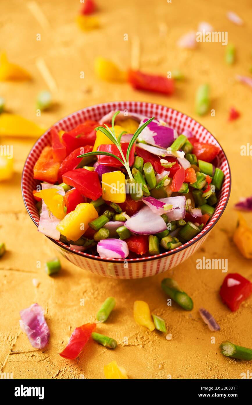 gros plan d'un bol avec un mélange de différents légumes crus hachés, tels que les asperges, l'oignon ou le poivron jaune et rouge, sur une surface dorée texturée Banque D'Images